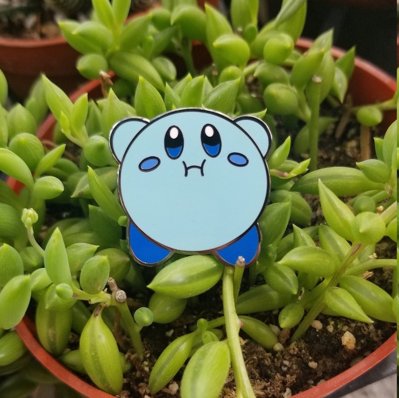 May include: A light blue enamel pin featuring a cartoon character with a sad expression. The character has large eyes, a small mouth, and a round body. The pin is set against a background of green succulent plants.