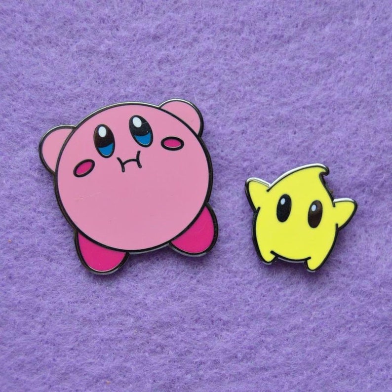 May include: Two enamel pins featuring characters from the video game Kirby. The first pin is pink and depicts Kirby with a surprised expression. The second pin is yellow and depicts a star-shaped character with a surprised expression.
