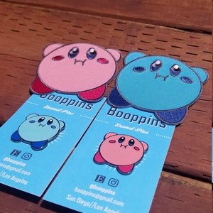 May include: Two enamel pins featuring a pink and a blue Kirby character. The pins are on blue cards with the text "Booppins Enamel Pins" and contact information for the seller.
