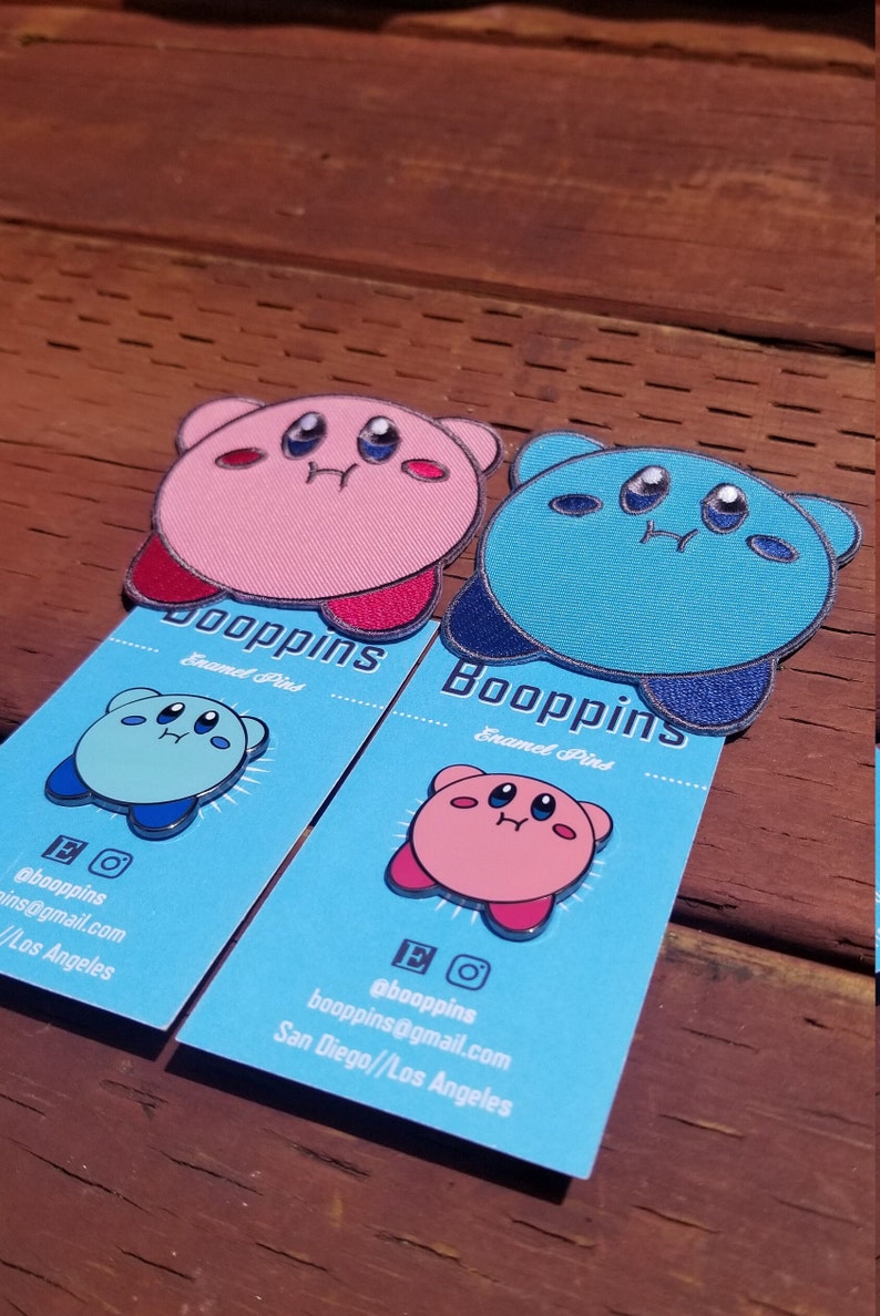 May include: Two enamel pins featuring a pink and a blue Kirby character. The pins are on blue cards with the text "Booppins Enamel Pins" and contact information for the seller.