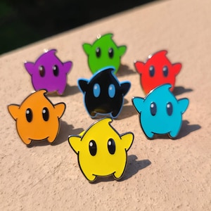 May include: A collection of seven colorful star-shaped enamel pins. The pins feature a variety of colors, including purple, green, red, blue, orange, black, and yellow. Each pin has two oval-shaped eyes.