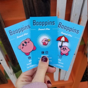 May include: Three blue cards with the text "Booppins Enamel Pins" and images of pink cartoon characters with a star, an umbrella, and a surprised expression. The cards are held in a hand.
