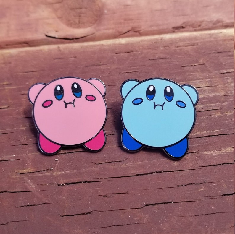 May include: Two enamel pins featuring a pink and a blue character with large eyes and a small mouth. The characters are round and have a simple design.