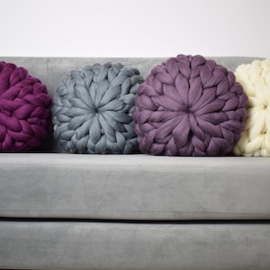 May include: Four round knitted throw pillows in various colors, including purple, gray, and white, are arranged on a gray couch.