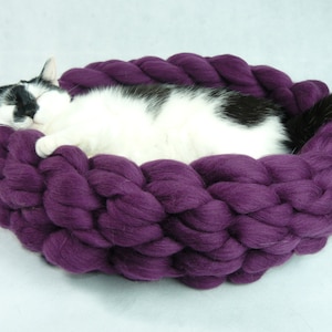 May include: A black and white cat sleeping in a purple knitted cat bed.