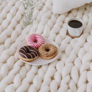 May include: Three donuts, one with chocolate frosting, one with pink and white frosting, and one with brown sprinkles, on a white plate with a cup of coffee and a small bouquet of white flowers on a white knitted blanket.