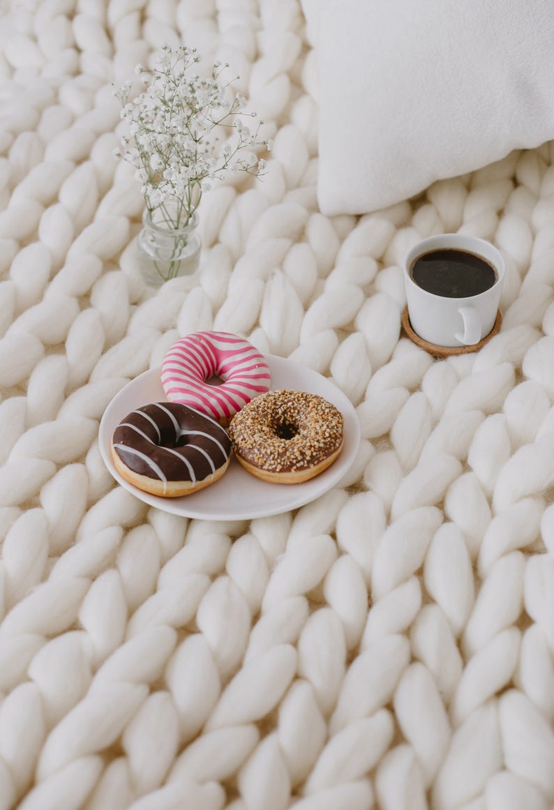 May include: Three donuts, one with chocolate frosting, one with pink and white frosting, and one with brown sprinkles, on a white plate with a cup of coffee and a small bouquet of white flowers on a white knitted blanket.