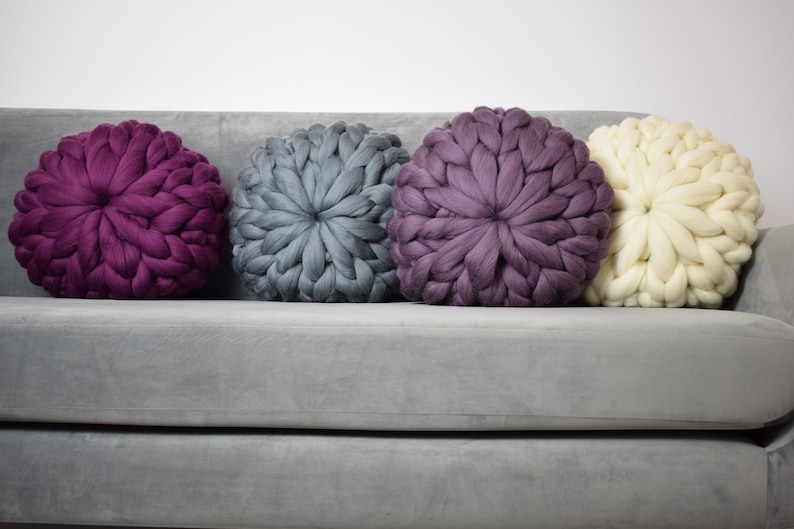 May include: Four round knitted throw pillows in various colors, including purple, gray, and white, are arranged on a gray couch.