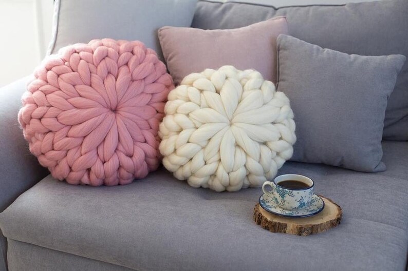 May include: Two large, round knitted throw pillows, one pink and one white, are on a gray couch. A small cup of coffee sits on a wooden coaster in front of the pillows.