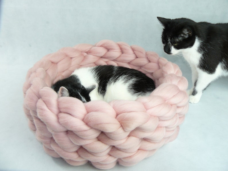 May include: A pink knitted cat bed with a black and white cat sleeping inside. The cat bed is made of thick, chunky yarn and has a soft, cozy texture.