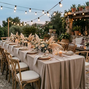 以下が含まれることがあります： 特別な日のために美しくセットされた屋外のダイニングテーブル。テーブルはベージュのテーブルクロスとランナーで覆われ、フラワーアレンジメント、キャンドル、食器で飾られています。上にはストリングライトが吊り下げられ、温かい雰囲気を作り出しています。