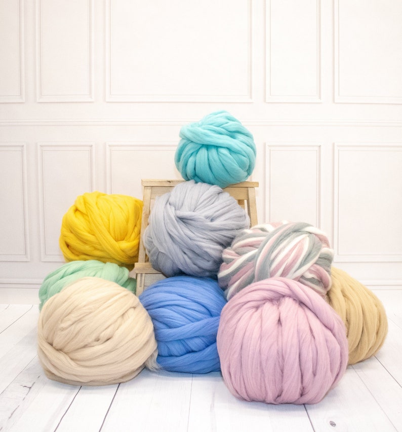 May include: A collection of eight large yarn balls in various colors, including yellow, blue, pink, green, and gray. The yarn balls are arranged on a wooden stool and a white surface.
