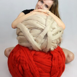 May include: A woman is sitting on a large red ball of yarn, holding a large beige ball of yarn in her arms.