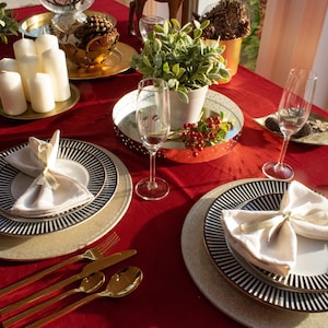 May include: A red tablecloth with two place settings, each with a white napkin, a black and white striped charger plate, a white dinner plate, and gold silverware. There are two champagne flutes and a small potted plant with green leaves on the table.