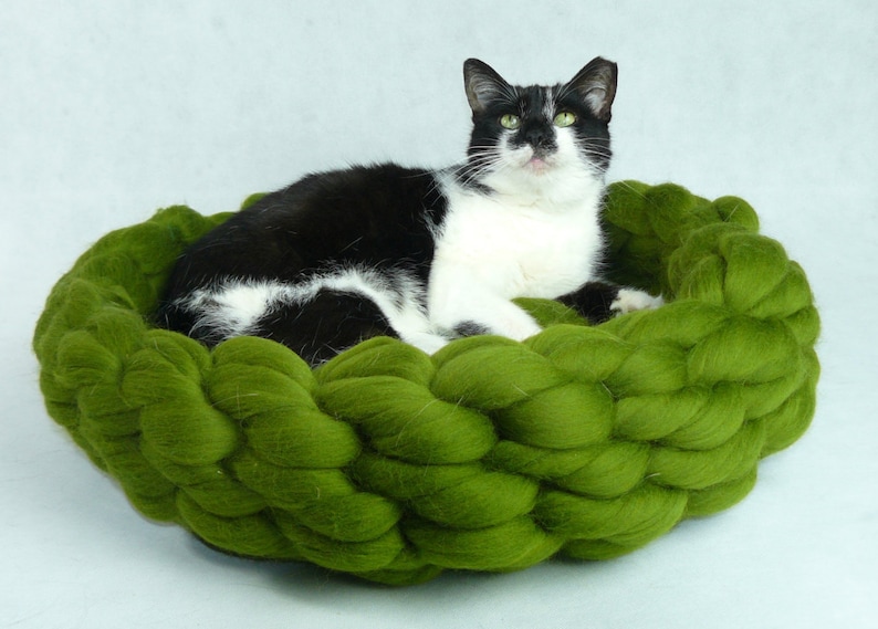 May include: A black and white cat resting in a green, woven pet bed. The bed is made of thick, braided yarn and has a round shape.