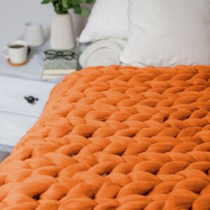 May include: A chunky knit throw blanket in a bright orange color. The blanket is made of thick yarn and has a soft, cozy texture. It is draped over a bed with white bedding and pillows.