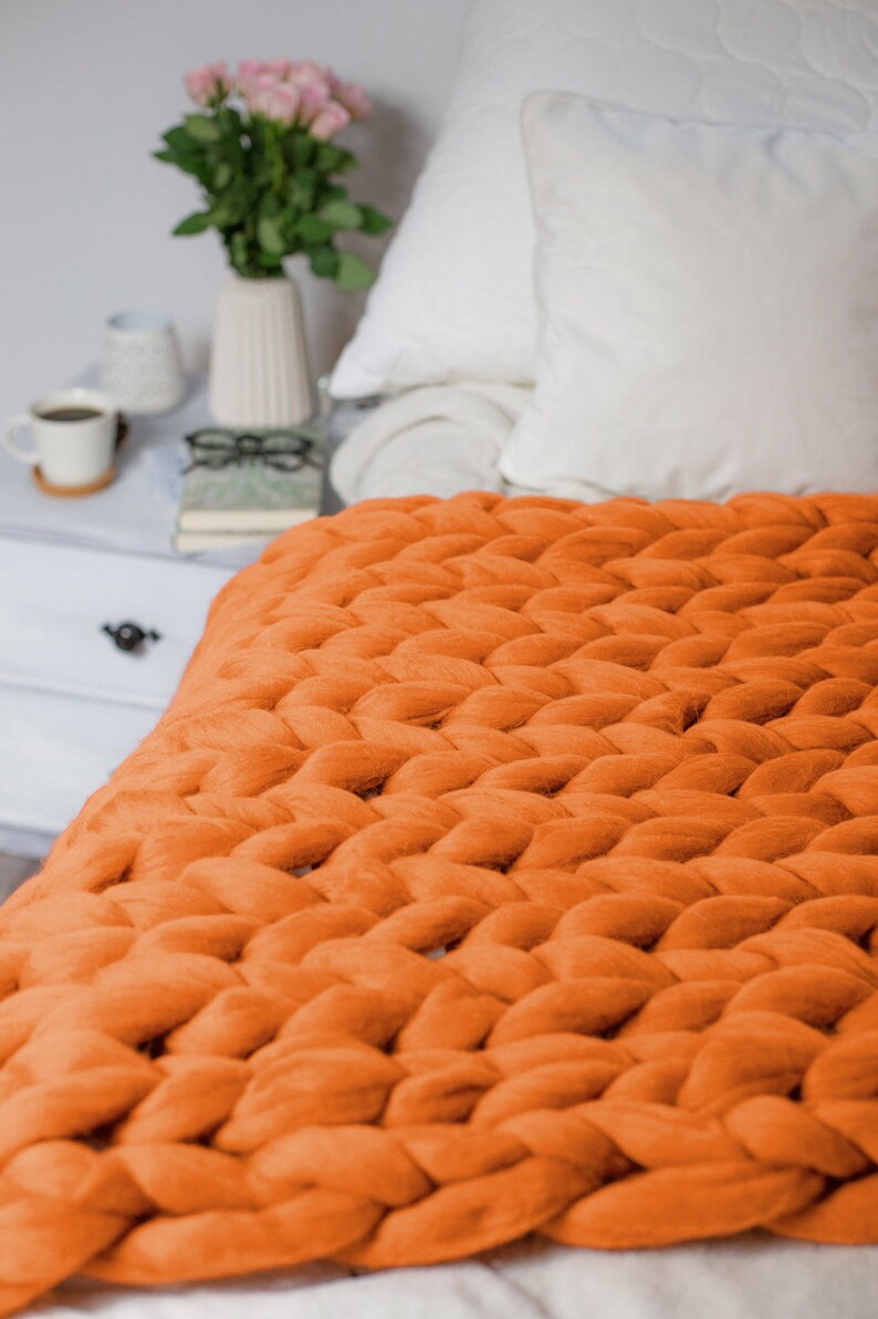May include: A chunky knit throw blanket in a bright orange color. The blanket is made of thick yarn and has a soft, cozy texture. It is draped over a bed with white bedding and pillows.