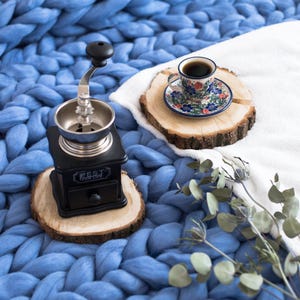 May include: A black manual coffee grinder with a silver top and a wooden base sits on a wooden coaster. A cup of coffee sits on a wooden coaster with a floral patterned saucer. The items are on a blue chunky knit blanket.
