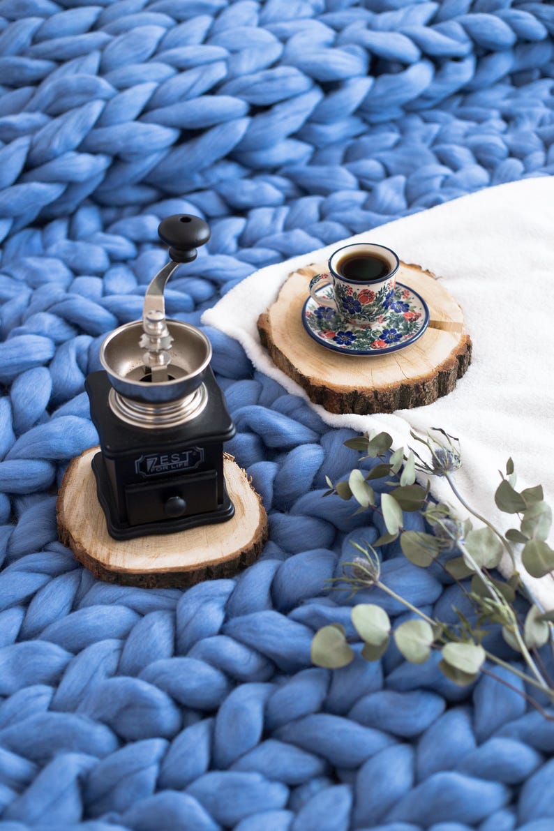 May include: A black manual coffee grinder with a silver top and a wooden base sits on a wooden coaster. A cup of coffee sits on a wooden coaster with a floral patterned saucer. The items are on a blue chunky knit blanket.