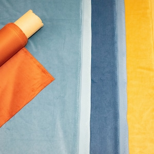 May include: A close-up of a variety of fabric swatches in different colors, including orange, blue, yellow, and gray. The swatches are arranged in a row, and a roll of orange fabric is lying on top of the blue swatch.