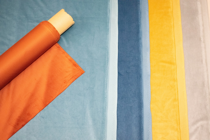 May include: A close-up of a variety of fabric swatches in different colors, including orange, blue, yellow, and gray. The swatches are arranged in a row, and a roll of orange fabric is lying on top of the blue swatch.