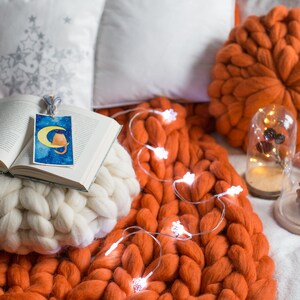 May include: A white knitted throw pillow with a book on top. The book has a bookmark with a cat illustration. The pillow is on a large orange knitted throw blanket with string lights.