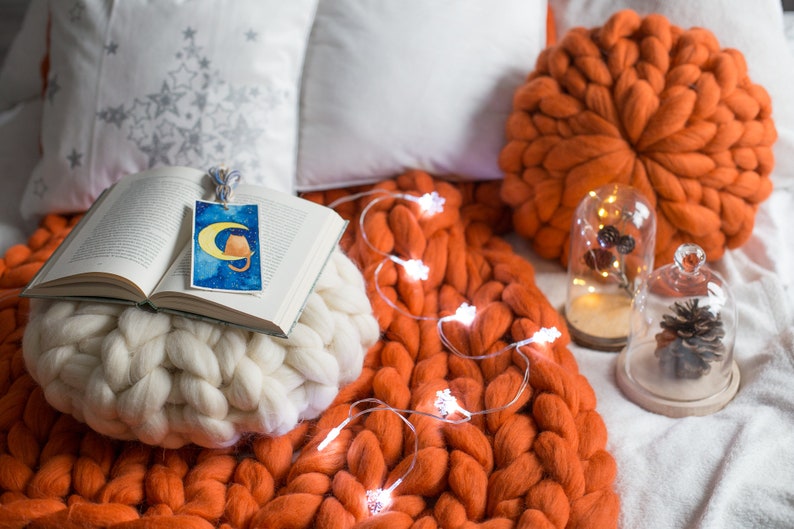 May include: A white knitted throw pillow with a book on top. The book has a bookmark with a cat illustration. The pillow is on a large orange knitted throw blanket with string lights.