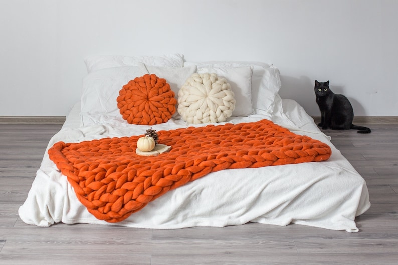 May include: A white bed with a chunky knit orange throw blanket and two round pillows, one white and one orange. A small white pumpkin sits on a wooden log on the blanket. A black cat sits on the floor in the background.