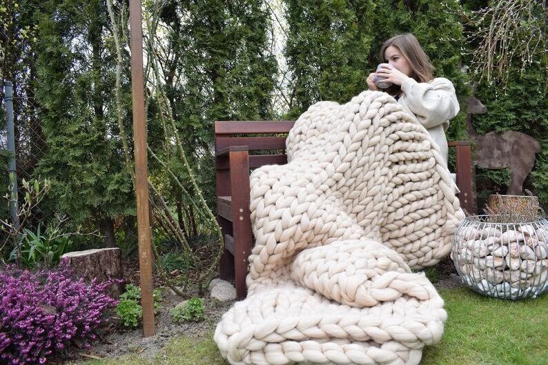 K&ouml;nnte beinhalten: Eine gro&szlig;e, grob gestrickte Wolldecke in Cremewei&szlig; liegt &uuml;ber einer Holzbank. Die Decke ist aus dickem Garn gefertigt und hat eine weiche, kuschelige Textur.