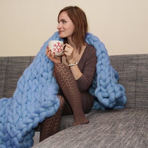 May include: A woman sits on a gray couch wrapped in a large, light blue knitted blanket. She is holding a white mug with a red snowflake design.