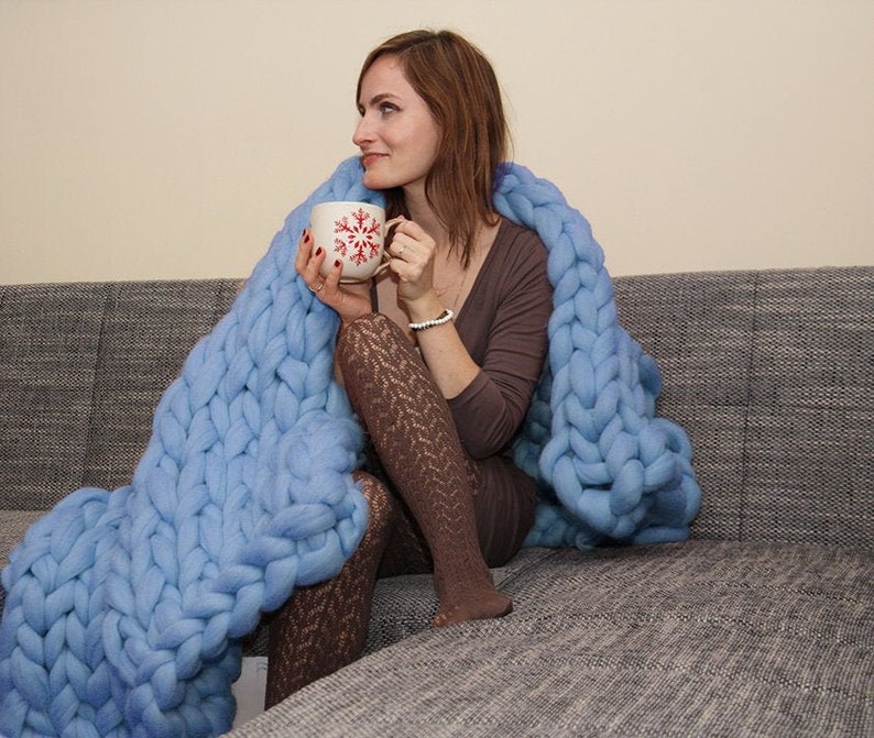 May include: A woman sits on a gray couch wrapped in a large, light blue knitted blanket. She is holding a white mug with a red snowflake design.