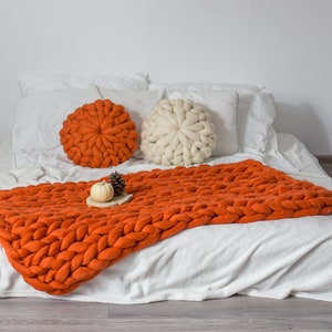 May include: A white bed with a chunky knit orange throw blanket and two round pillows, one orange and one white. The bed is on a wood floor.