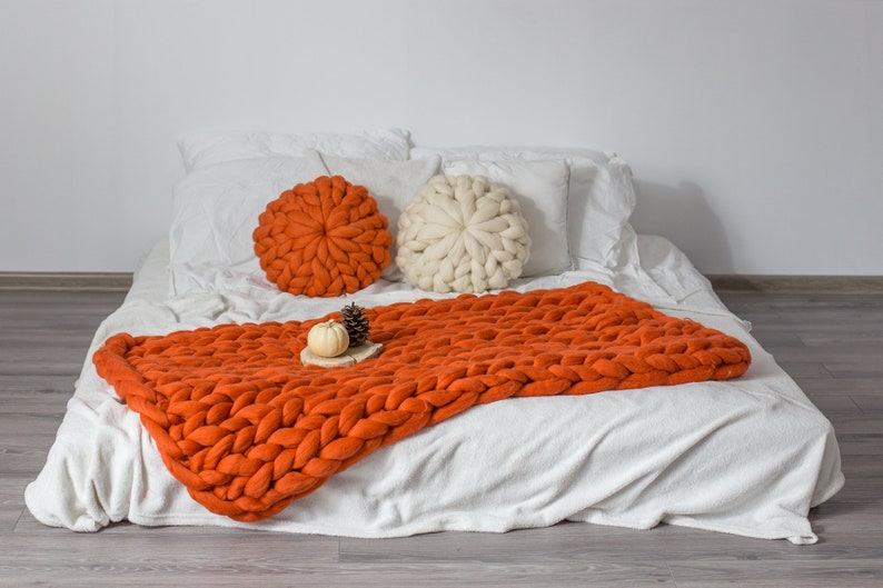 May include: A white bed with a chunky knit orange throw blanket and two round pillows, one orange and one white. The bed is on a wood floor.