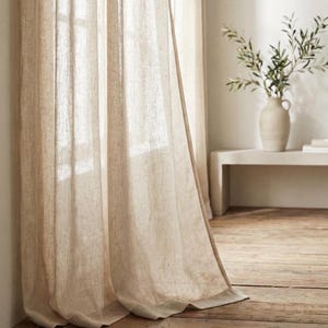 Puede incluir: Cortinas de lino beige cuelgan frente a una ventana, dejando pasar la luz del sol. Las cortinas tienen un aspecto texturizado y una caída suave. Un jarrón blanco con ramas verdes se encuentra sobre una estantería blanca en el fondo.