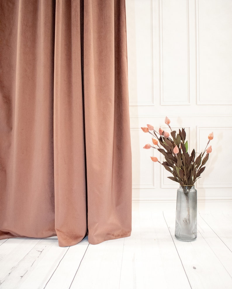 May include: A vase with dried flowers in front of a brown curtain. The vase is clear glass with a ribbed texture. The flowers are a light pink color.