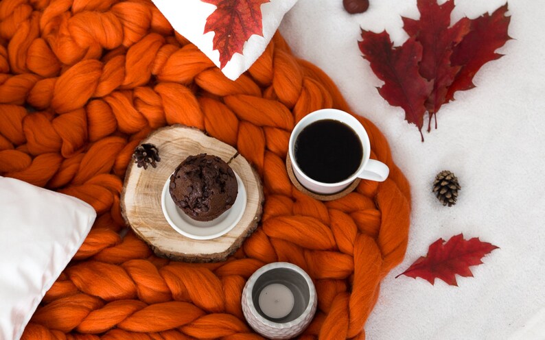 May include: A cozy autumn scene with a cup of coffee, a chocolate muffin, and a chunky knit orange blanket. Red fall leaves and pine cones are scattered around the blanket.