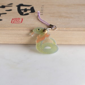 Puede incluir: Un amuleto de serpiente verde y naranja con una campana en una cuerda morada. El amuleto está hecho de plástico transparente y tiene un efecto 3D.