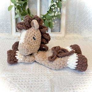 May include: A handmade, plush crochet horse in shades of brown, tan, and white. The horse is lying down, with a fluffy mane and tail. The toy is soft and cuddly, perfect for a child's bedroom or nursery.