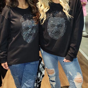 May include: Two women wearing black long-sleeved shirts with a rhinestone skull design. The women are wearing blue jeans and silver glitter sneakers.