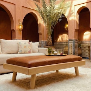 May include: A cream-colored sofa with decorative pillows and a brown leather ottoman on a light wood frame. The room has arched doorways, hanging lanterns, and a white rug. The walls are painted in a warm terracotta color.