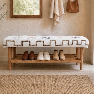 May include: A rectangular wooden bench with four tapered legs and a lower storage shelf. The seat is upholstered in white fabric featuring a brown geometric border pattern and small, colorful embroidered accents. Three pairs of shoes sit on the shelf.