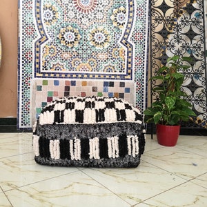 May include: Black and white patterned pouf ottoman with a fluffy texture. The pouf is made of woven wool and has a square shape.