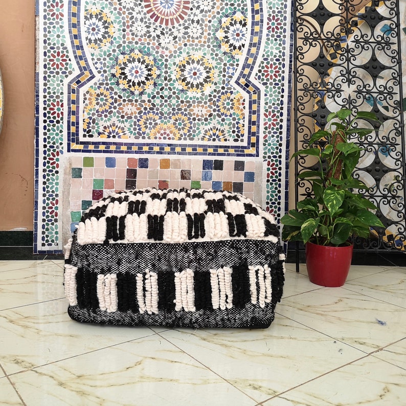 May include: Black and white patterned pouf ottoman with a fluffy texture. The pouf is made of woven wool and has a square shape.