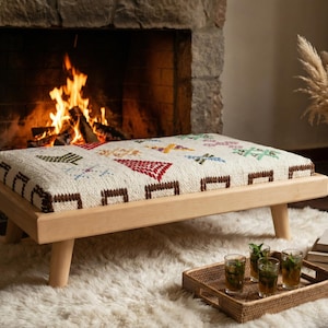 May include: A rectangular wooden ottoman with a patterned, off-white cushion. The cushion features colorful geometric designs. The ottoman sits on a fluffy white rug, with a tray of drinks and a vase of dried flowers nearby. A fireplace with a burning fire is in the background.
