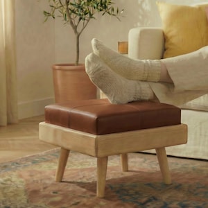 May include: A square ottoman featuring a smooth, dark brown leather top cushion and a light-toned wooden base with four tapered legs. The piece is designed with a clean, low-profile structure, showcasing a contrast between the leather and wood.