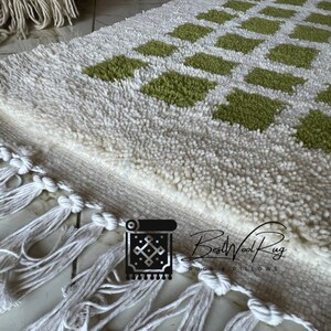 May include: White and green wool rug with a geometric pattern and fringe. The rug is made by BestWoolRug.