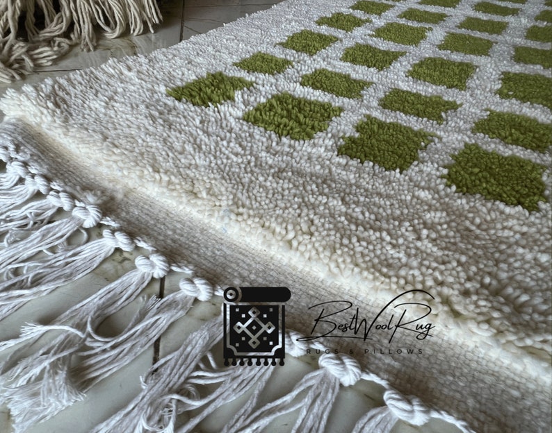 May include: White and green wool rug with a geometric pattern and fringe. The rug is made by BestWoolRug.