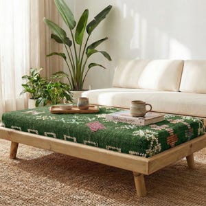 May include: A living room scene featuring a low wooden coffee table with a green patterned cushion, a beige sofa, and a woven rug. The room is bright with natural light, and includes plants and decorative items.