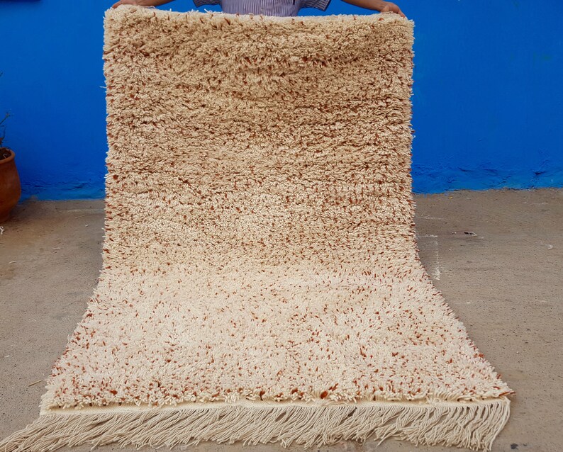 K&ouml;nnte beinhalten: Ein cremefarbener Teppich mit einem dezenten braunen Fleckenmuster. Der Teppich hat einen Fransenrand und ist aus einem weichen, flauschigen Material gefertigt.