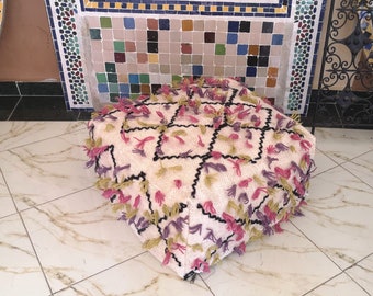 Puede incluir: Un puf blanco con un patrón geométrico en negro, rosa y verde. El puf tiene un borde de flecos y está hecho de un material suave y esponjoso.